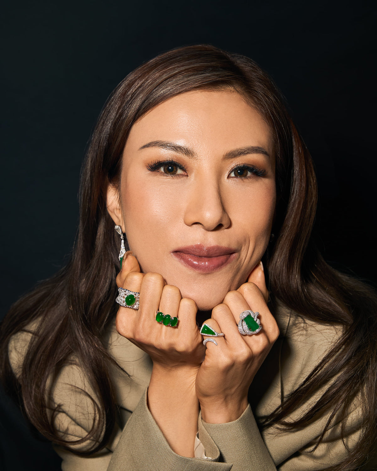 Editorial portrait of model with hand resting against her head, looking directly at the camera, showcasing four jadeite and diamond rings worn across the knuckles — The Oval Mosaic Ring, The Trilogy Oval Cabochon Ring, The Orbit Ring in triangular bypass setting, and The Cylinder Knot Ring — with only the diamond briolette of The Cylinder Drop Earrings visible, demonstrating the pieces' capacity for layered daily wear.