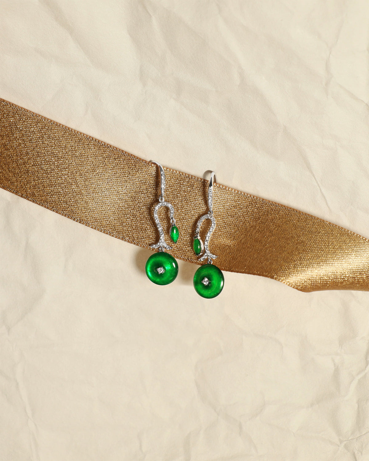 Bright green jadeite drop earrings with a single pavé diamond arc, small marquise stone at the top, and a round disc at the base, set against a white background accented with a gold ribbon, front view.