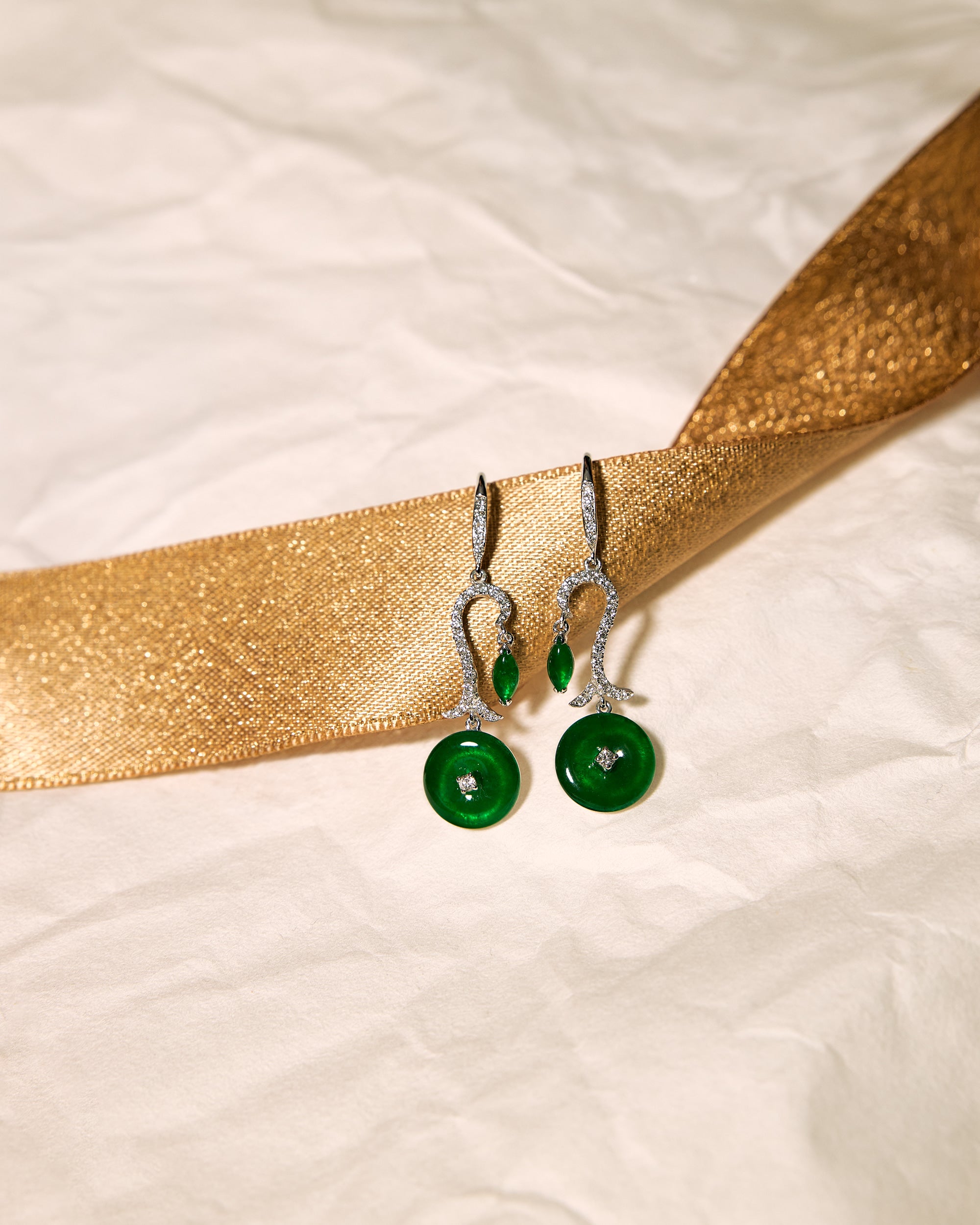 Bright green jadeite drop earrings with a single pavé diamond arc, small marquise stone at the top, and a round disc at the base, set against a white background accented with a gold ribbon, front view.