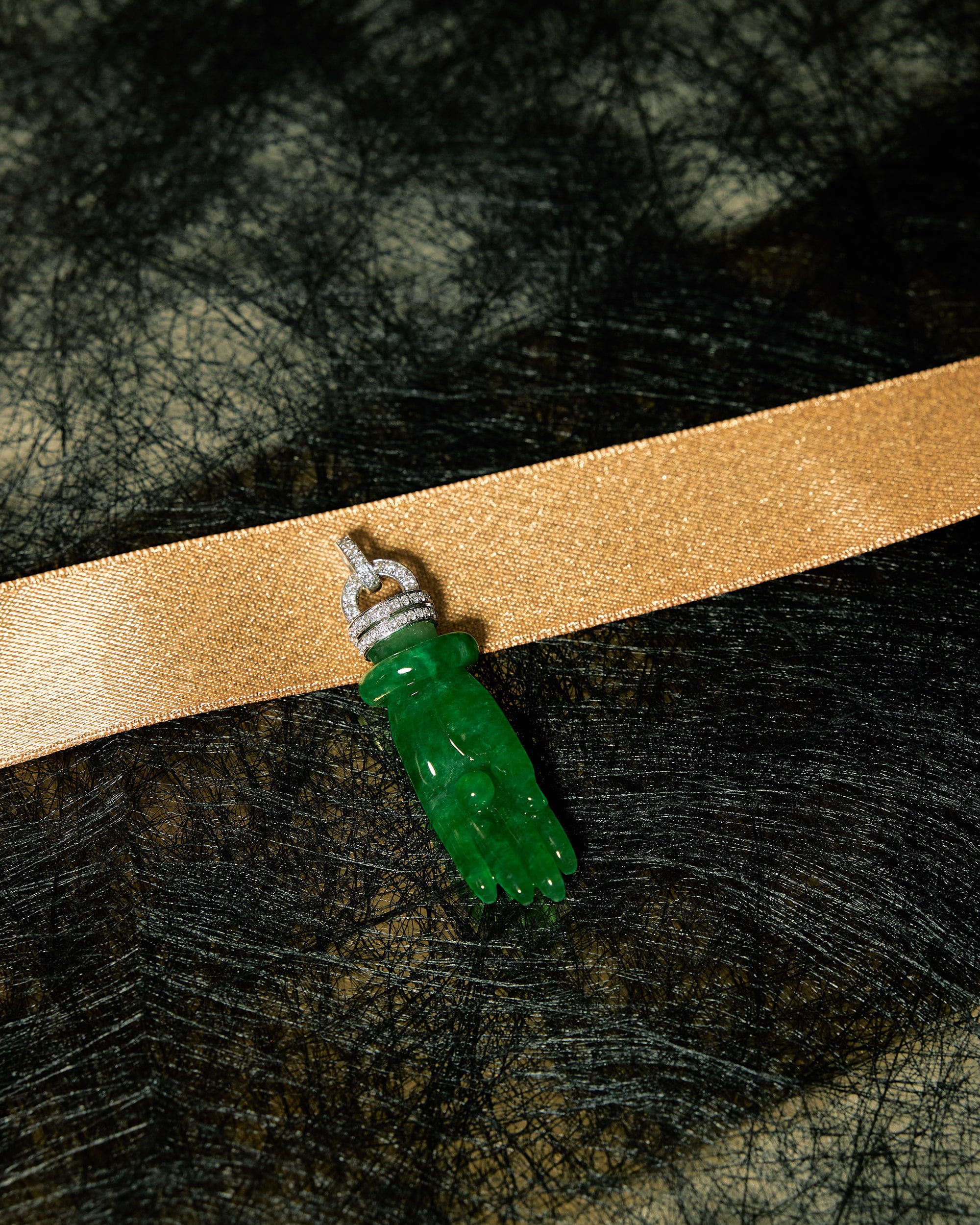 Carved bright green jadeite Buddha's palm pendant with articulated bangle and diamond pavé rows set against a black background accented with a gold ribbon, front view.