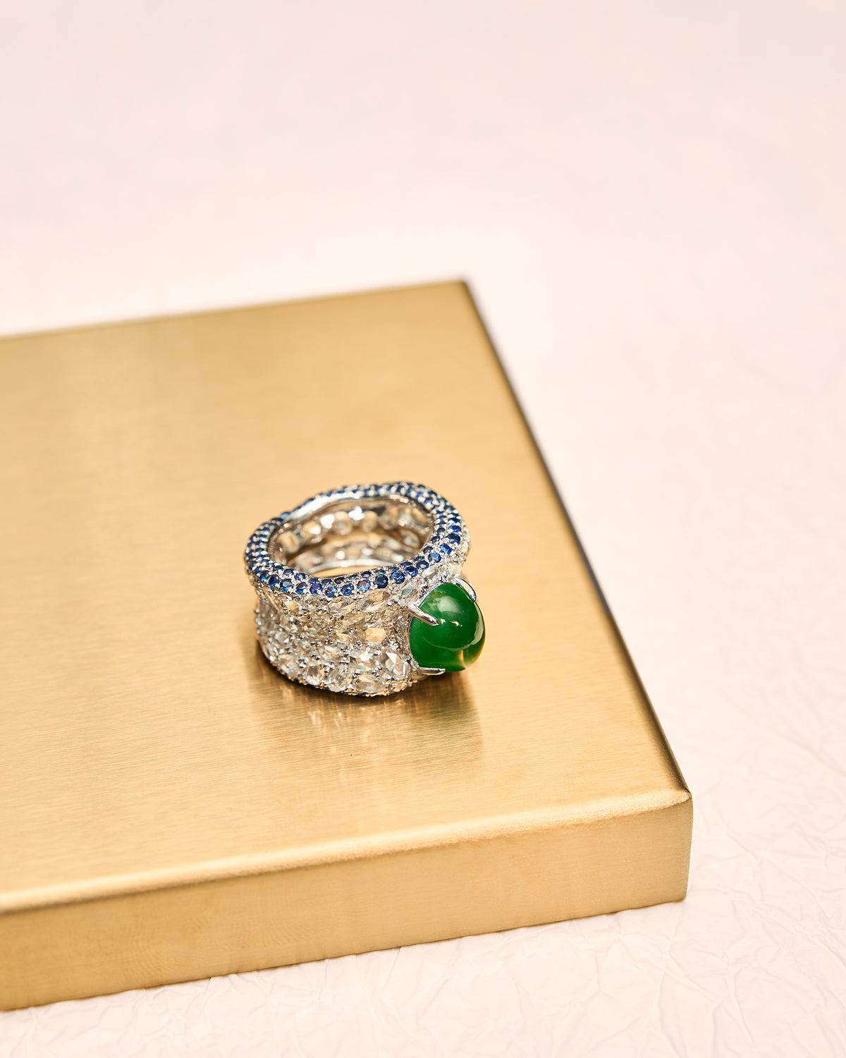 Top-down view of The Oval Mosaic Ring on a gold tray, revealing the interior of the band alongside the intense green oval jadeite cabochon, wide rose-cut diamond pavé, and sapphire borders.