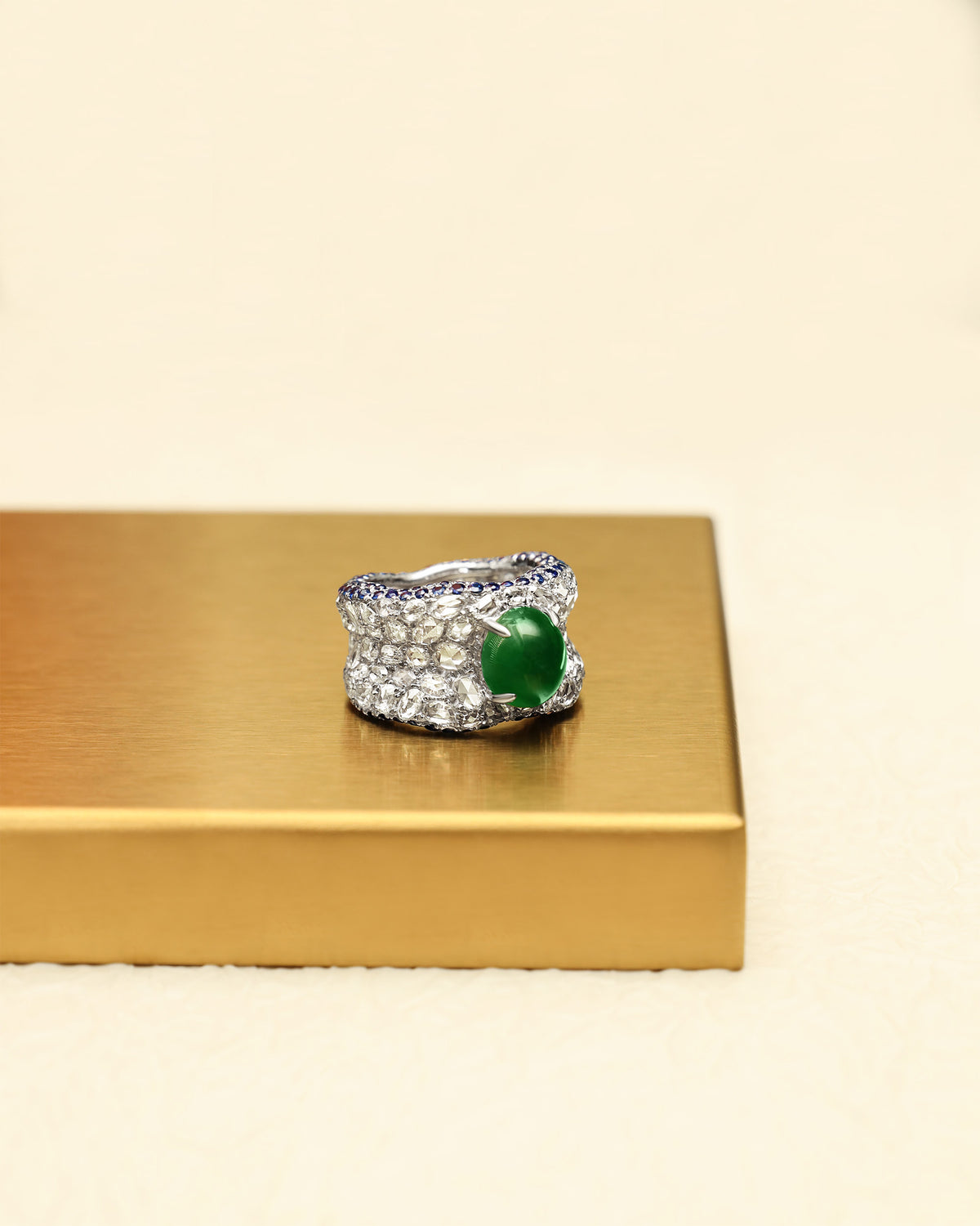Three-quarter front view of The Oval Mosaic Ring on a gold tray, showing the intense green oval jadeite cabochon with wide rose-cut diamond pavé and sapphire borders.
