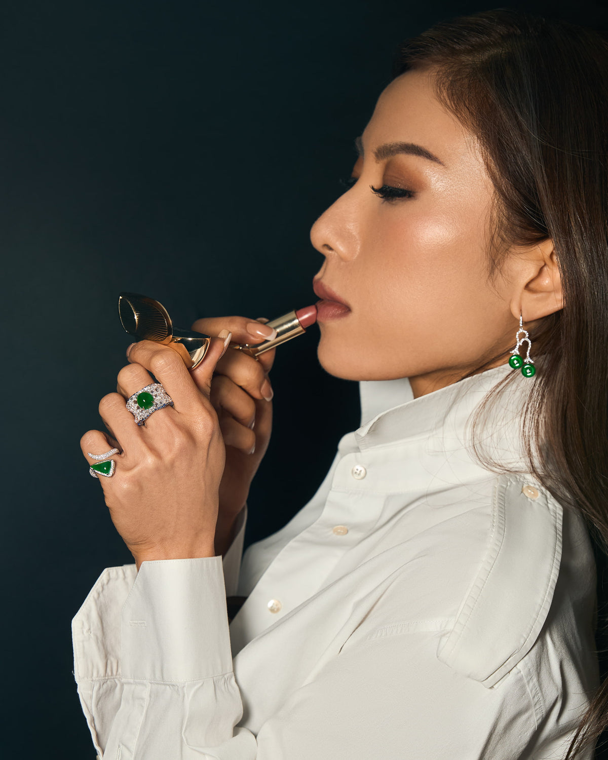 Editorial side profile of model applying lipstick, wearing The Oval Mosaic Ring and The Orbit Ring in triangular bypass setting on her fingers, and The Twin Arc Drop Earrings visible against the profile silhouette, demonstrating the pieces' architectural restraint and suitability for daily ritual.