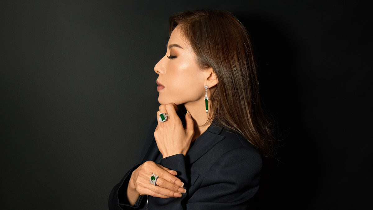 Editorial side profile of model in black blazer against a clean black studio background, left hand resting thoughtfully at chin, right hand holding left elbow, wearing The Oval Mosaic Ring on right hand and The Cylinder Knot Ring on left hand, with The Cylinder Drop Earrings visible, face turned toward the left in a contemplative pose, demonstrating the pieces' architectural restraint and suitability for daily wear.