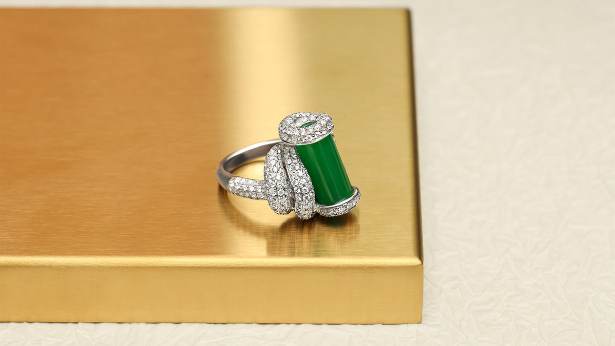 The Cylinder Knot Ring featuring an intense green jadeite cylinder with pavé diamond rope knot setting on the shank, displayed on a gold tray, side view showing the upright cylinder profile and the diamond knot structure wrapping around the band.