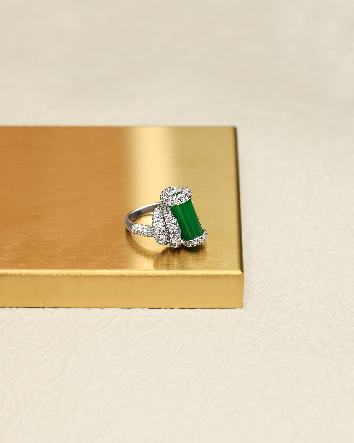 The Cylinder Knot Ring featuring an intense green jadeite cylinder with pavé diamond rope knot setting on the shank, displayed on a gold tray, side view showing the upright cylinder profile and the diamond knot structure wrapping around the band.