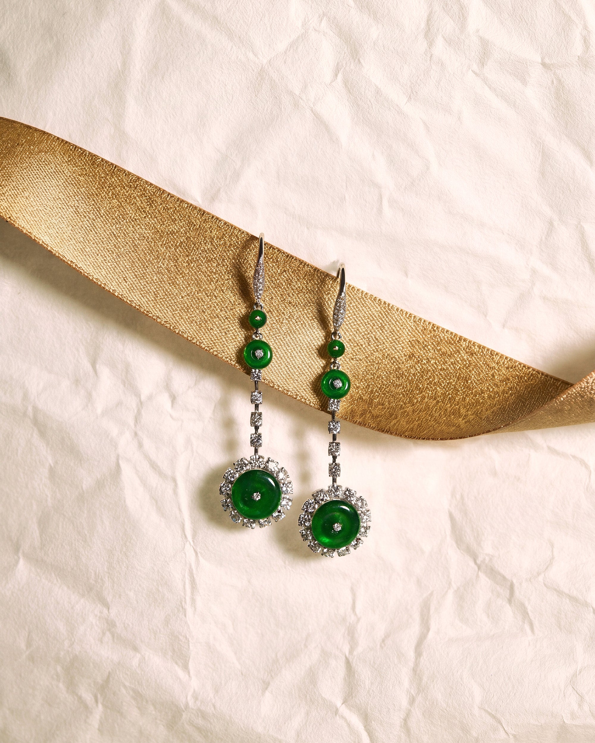 Bright green jadeite drop earrings featuring three graduated discs with a diamond-set finale, highlighting scale progression, set against a white background accented with a gold ribbon, front view.