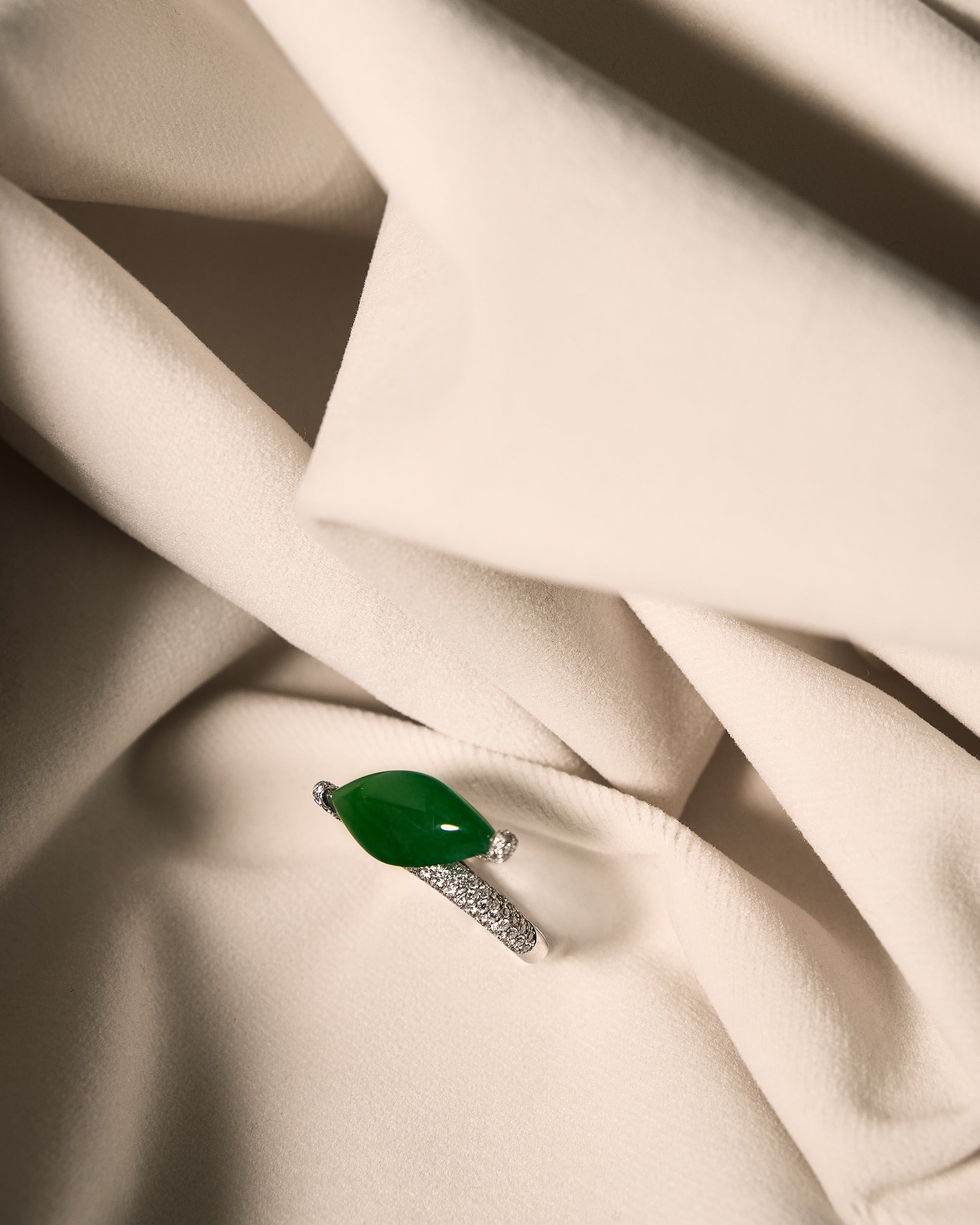 Intense green marquise jadeite ring with east-west bypass pavé diamond band on ivory cloth, front view showing full unobstructed stone face.