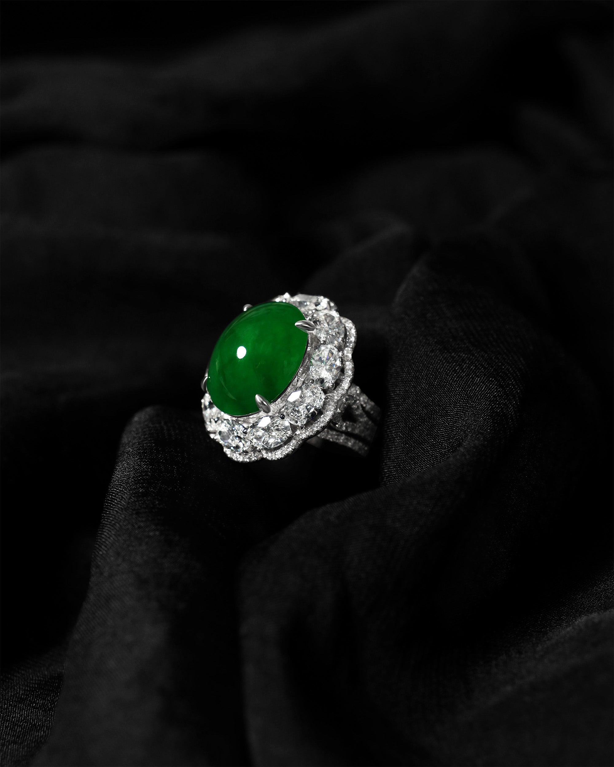Intense green oval jadeite cabochon ring framed by ten matched oval diamonds and a three-quarter pavé band on a black cloth, three quarter view.
