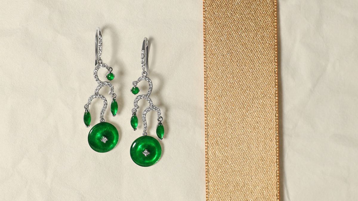 Front view of The Tiered Arc Drop Earrings showing three descending pavé arcs with a round jadeite at the top, marquise-shaped jadeite in the middle, and a huaigu disc at the base, set against a white background accented with a gold ribbon.