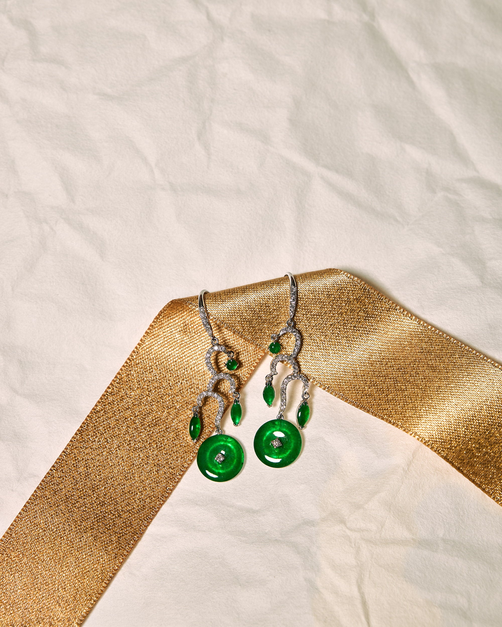 Front view of The Tiered Arc Drop Earrings showing three descending pavé arcs with a round jadeite at the top, marquise-shaped jadeite in the middle, and a huaigu disc at the base, set against a white background accented with a gold ribbon.